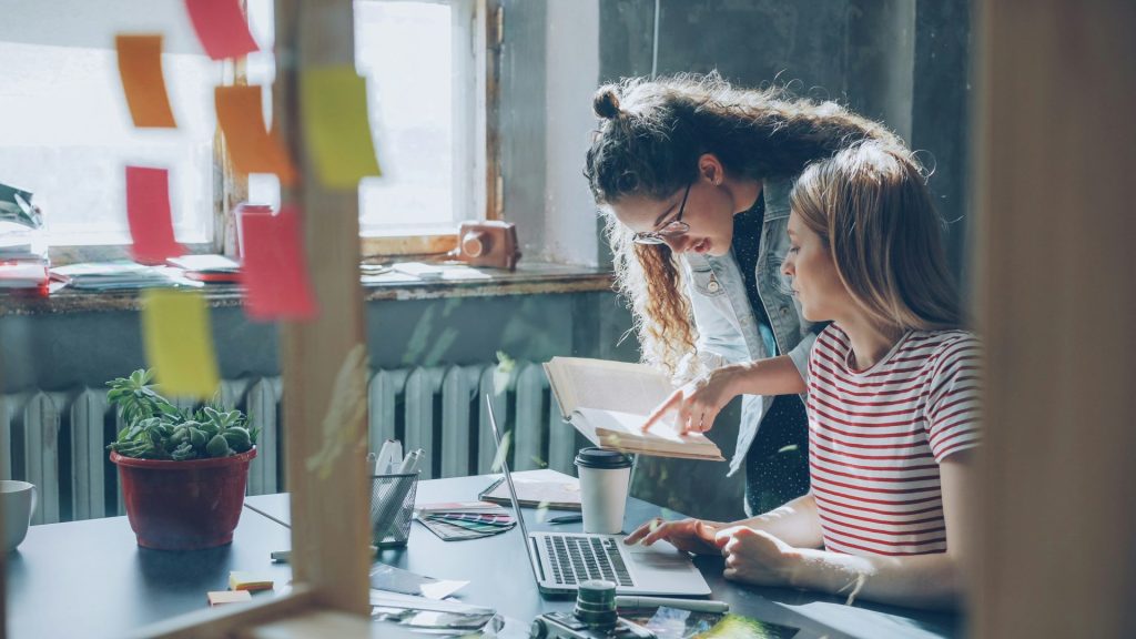 Two women collaborate over a laptop in an office.